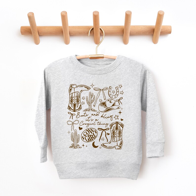 May include: A light grey sweatshirt with a brown graphic design featuring cowboy boots, a hat, cactus, and the text "Boots and Bling it's a Cowgirl thing". The sweatshirt is hanging on a wooden hanger.