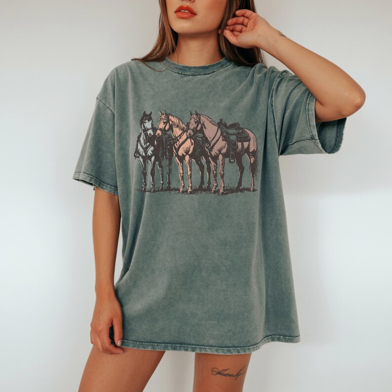 May include: A faded green t-shirt featuring a vintage-style illustration of four horses. The horses are depicted in shades of brown, tan, and gray, with saddles and tack visible on some. The shirt has a relaxed fit and short sleeves.