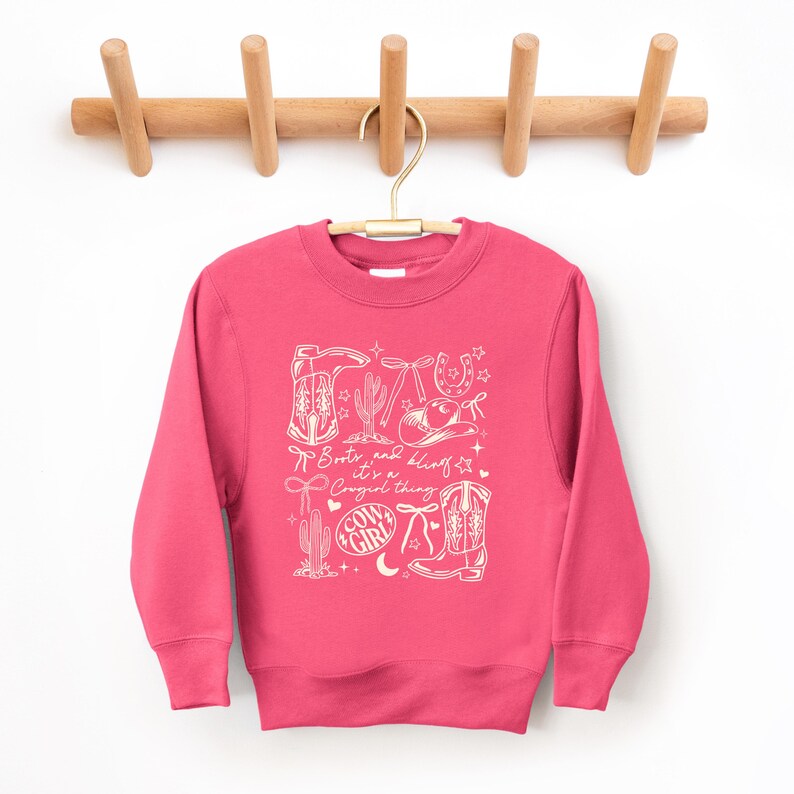 May include: A cerise sweatshirt with a white graphic design featuring cowboy boots, a cactus, a horseshoe, and a cowboy hat. The text "Boots and Bling, it's a Cowgirl thing" is also included in the design.