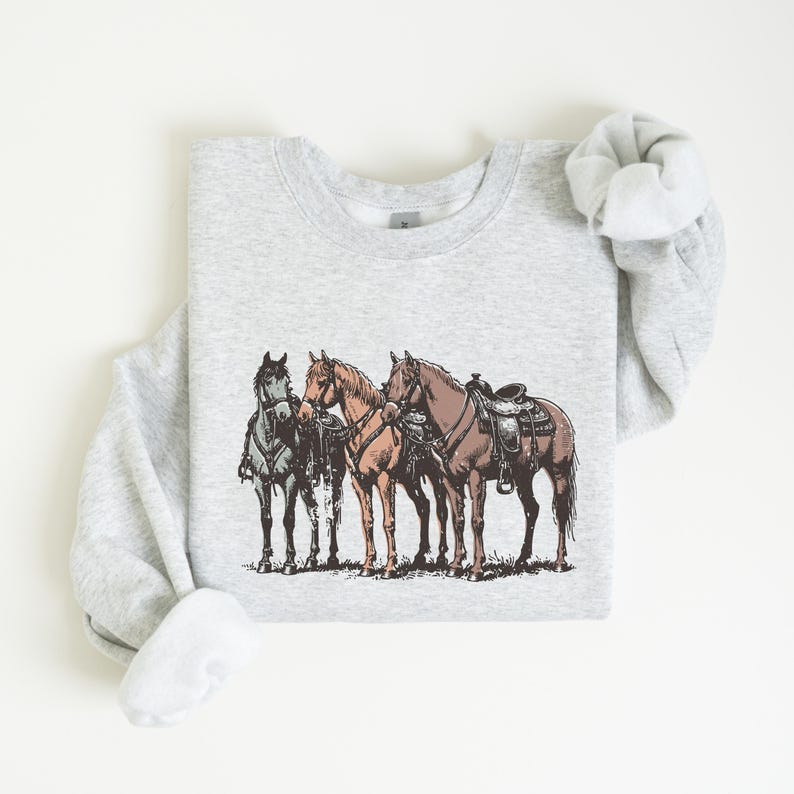 May include: A light gray sweatshirt featuring a detailed illustration of three horses. The horses are depicted in shades of brown, tan, and gray, with one saddled. The design has a vintage, hand-drawn aesthetic, suggesting a Western or equestrian theme.