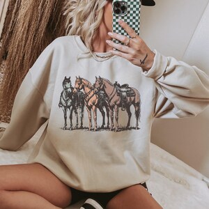 May include: Cream-colored sweatshirt featuring a graphic of three horses in shades of brown, tan, and gray. The horses are standing side-by-side, with one saddled. The design is printed on the front of the long-sleeved top.