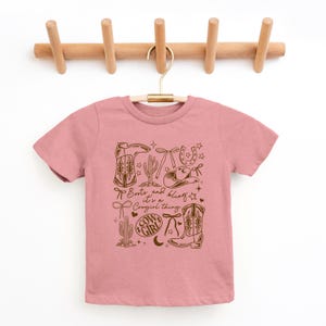 May include: A pink t-shirt with a brown graphic design featuring cowboy boots, a hat, a horseshoe, and cacti. The text "Boots and bling, it's a Cowgirl thing" is printed on the shirt.