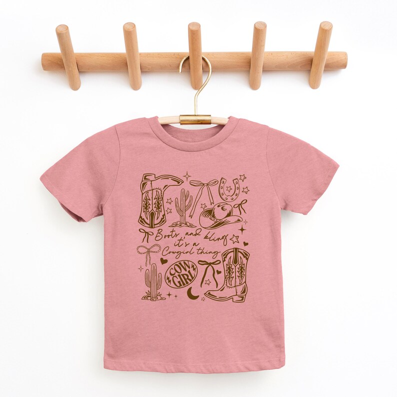 May include: A pink t-shirt with a brown graphic design featuring cowboy boots, a hat, a horseshoe, and cacti. The text "Boots and bling, it's a Cowgirl thing" is printed on the shirt.