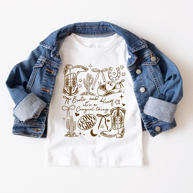 May include: White t-shirt with a brown graphic design featuring cowboy boots, a horseshoe, cactus, and the text "Boots and bling it's a Cowgirl thing." The shirt is paired with a blue denim jacket.