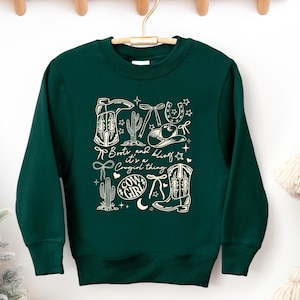 May include: A dark green sweatshirt with a white graphic design featuring cowboy boots, a cowboy hat, cacti, and the text "Boots and bling it's a Cowgirl thing". The design includes stars, a horseshoe, and a bow.
