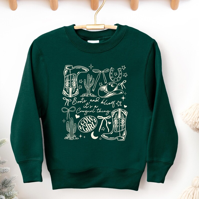 May include: A dark green sweatshirt with a white graphic design featuring cowboy boots, a cowboy hat, cacti, and the text "Boots and bling it's a Cowgirl thing". The design includes stars, a horseshoe, and a bow.