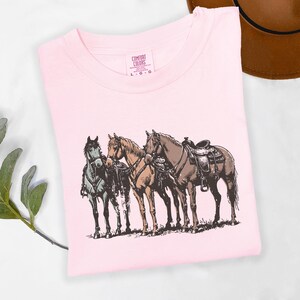 May include: A light pink Comfort Colors t-shirt featuring a graphic of three horses. The horses are in shades of brown, tan, and grey, with saddles on two of them. The shirt is folded and has a tag in the upper center.