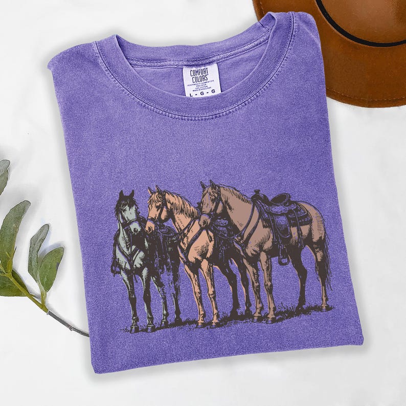 May include: A purple Comfort Colors t-shirt featuring a graphic of three horses with saddles. The horses are in shades of brown, tan, and green. The shirt has a crew neck and the Comfort Colors logo is visible.