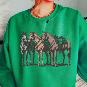 May include: Green sweatshirt featuring a vintage-style illustration of three horses. The horses are depicted in shades of brown, tan, and gray, with saddles and bridles. The sweatshirt is a solid green color, creating a contrast with the detailed graphic.