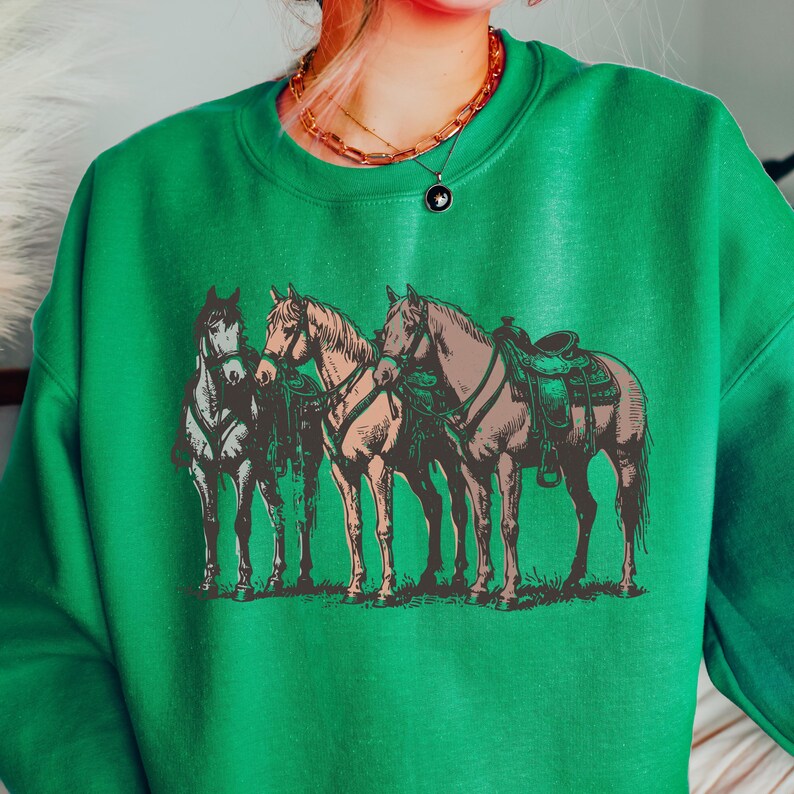 May include: Green sweatshirt featuring a vintage-style illustration of three horses. The horses are depicted in shades of brown, tan, and gray, with saddles and bridles. The sweatshirt is a solid green color, creating a contrast with the detailed graphic.