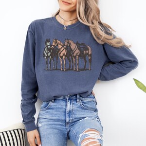 May include: A navy blue long-sleeve sweatshirt featuring a graphic of four horses. The horses are in shades of brown, gray, and tan, with saddles on two of them. The sweatshirt is made of a soft material.