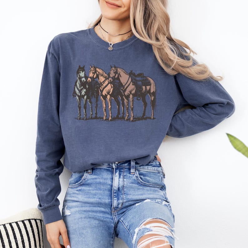 May include: A navy blue long-sleeve sweatshirt featuring a graphic of four horses. The horses are in shades of brown, gray, and tan, with saddles on two of them. The sweatshirt is made of a soft material.