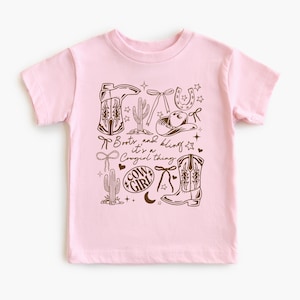 May include: A pale pink t-shirt with a brown graphic design. The design includes cowboy boots, a hat, a horseshoe, and cacti. The text "Boots and bling it's a Cowgirl thing" is also included.