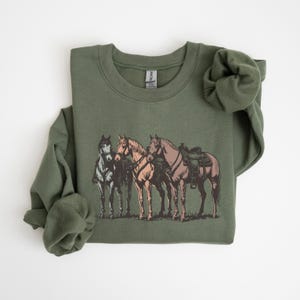 May include: Olive green sweatshirt featuring a graphic of three horses. The horses are depicted in shades of brown, tan, and gray, with saddles on two of them. The design is centered on the front of the sweatshirt.