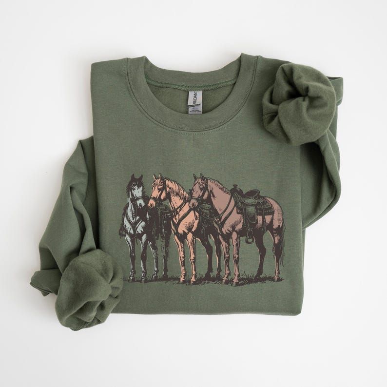 May include: Olive green sweatshirt featuring a graphic of three horses. The horses are depicted in shades of brown, tan, and gray, with saddles on two of them. The design is centered on the front of the sweatshirt.