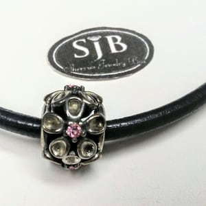 May include: A silver bead with a floral design and pink accents is strung on a black leather cord. The bead has a detailed pattern with clear and pink stones. The background includes a logo that reads "SJB Catherine's Jewelry Box."