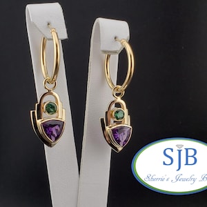 May include: Gold hoop earrings with a dangling charm featuring a purple triangular gemstone and a smaller green gemstone. The charm is set in a geometric design.