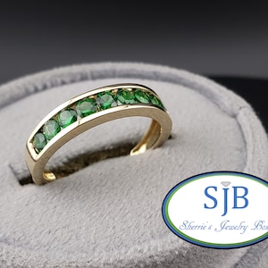Puede incluir: Un anillo de banda de oro con una fila de piedras preciosas verdes. El anillo se muestra sobre una superficie de terciopelo gris.