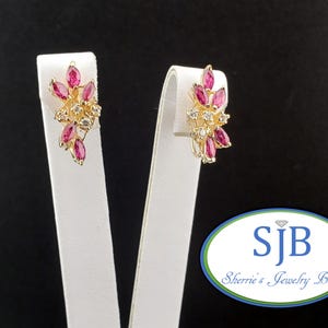 May include: A pair of gold-toned stud earrings. Each earring features a cluster of marquise-cut, ruby-colored stones and small, clear stones. The earrings are displayed on white stands against a black background. The earrings are from Sherrie's Jewelry Box.