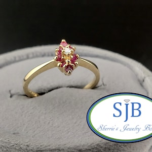 May include: A gold ring with a cluster of red gemstones and a single white gemstone at the center. The ring is set on a gray surface. The ring is from Sherrie's Jewelry Box.