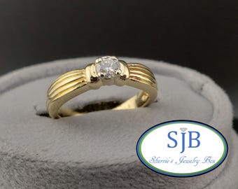 May include: A gold ring with a central diamond, set in a ribbed band. The ring is displayed on a gray velvet surface. The ring is from Sherrie's Jewelry Box.