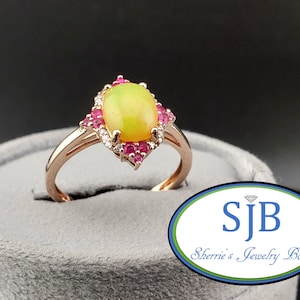 May include: A rose gold ring featuring a central oval cabochon yellow-green opal, surrounded by a halo of small round pink rubies and tiny clear accent stones. The ring has a split shank design and is displayed in a grey velvet ring box.