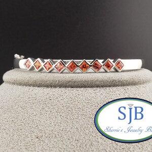 May include: A silver bangle bracelet featuring a line of square, orange gemstones. The bracelet is presented on a grey surface against a dark backdrop. The image includes the text "SJB Sherrie's Jewelry Box".