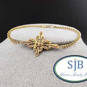 May include: A gold-toned bracelet featuring a central filigree cross-shaped ornament with intricate wirework and small spherical accents. The bracelet is constructed with a delicate chain and a simple hook clasp, showcasing a detailed metal design.