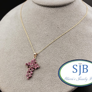 Pode incluir: Um colar dourado com um pendente em forma de cruz. A cruz é adornada com numerosas pedras preciosas pequenas, ovais e de cor vermelho escuro. O colar é exibido sobre uma superfície cinza. O pendente tem cerca de 3,8 cm de comprimento.