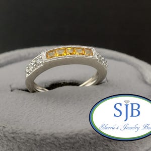 May include: A silver ring with five square, yellow gemstones set in a row. The ring has a detailed design with small, clear stones on the sides. The ring is displayed on a gray surface.