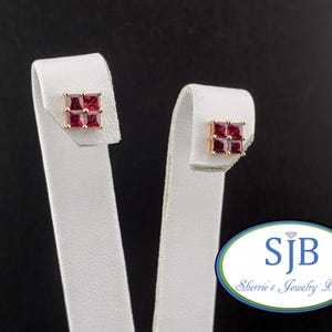 May include: Gold-tone square stud earrings, each featuring four square-cut, deep red gemstones. The earrings are displayed on white jewelry display stands against a black background. The logo "SJB Sherrie's Jewelry Box" is visible.