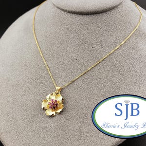 以下が含まれることがあります： 金色のネックレス、花のペンダント付き。ペンダントは花びらのデザインで、中央に小さな赤い宝石のクラスターがあります。ネックレスは灰色のマネキンに展示されています。