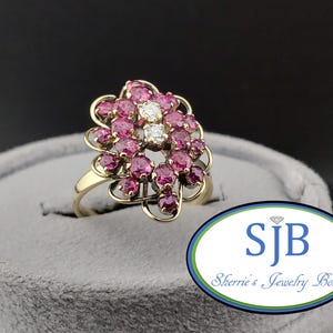 May include: A gold ring featuring a cluster of round, deep pink gemstones arranged in a floral-like pattern, with two clear, round, white stones set vertically in the center. The metal band is smooth and yellow-toned, supporting the ornate setting.
