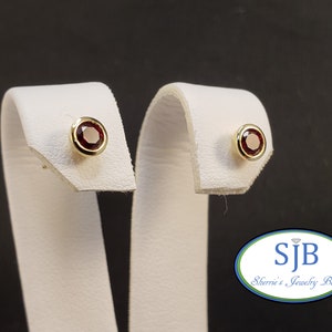 May include: A pair of gold stud earrings with red gemstones. The earrings are on a white display stand.