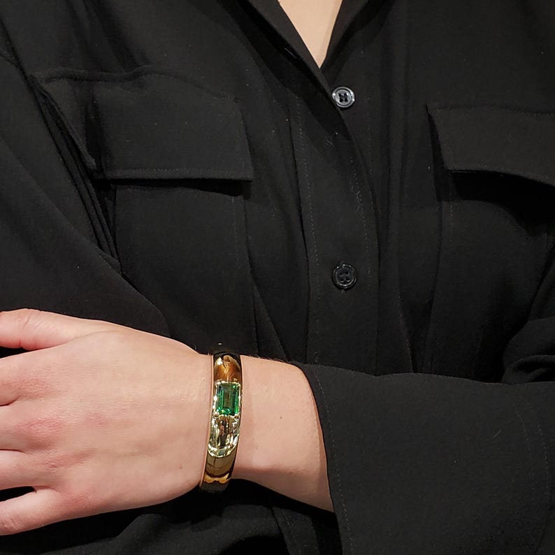 Może przedstawiać: Złota bransoletka typu bangle z zielonym szmaragdem osadzonym w środku.