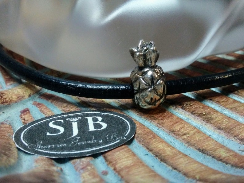 May include: A silver pineapple charm on a black leather cord. The charm is detailed with a textured pineapple design. The charm is on a wooden surface with a blue and brown striped pattern. The charm is labeled "SJB" for "Sherries Jewelry Box".