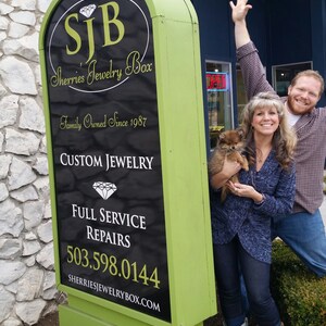 Pu&ograve; includere: Un cartello verde e nero con il testo "SJB Sherries Jewelry Box" e "Family Owned Since 1987". Il cartello include anche il testo "Custom Jewelry", "Full Service Repairs" e un numero di telefono. Una coppia e un piccolo cane stanno in piedi davanti al cartello.