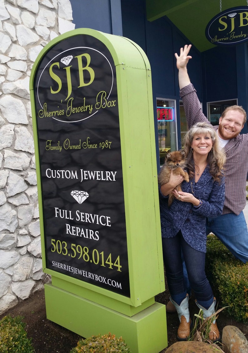 Pu&ograve; includere: Un cartello verde e nero con il testo "SJB Sherries Jewelry Box" e "Family Owned Since 1987". Il cartello include anche il testo "Custom Jewelry", "Full Service Repairs" e un numero di telefono. Una coppia e un piccolo cane stanno in piedi davanti al cartello.