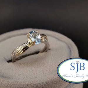 May include: A gold and silver ring with a light blue oval gemstone and small diamonds on the band. The ring is in a gray ring box.  SJB Sherrie's Jewelry Box.