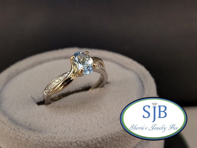 May include: A gold and silver ring with a light blue oval gemstone and small diamonds on the band. The ring is in a gray ring box.  SJB Sherrie's Jewelry Box.
