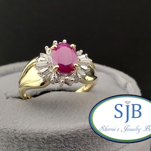 May include: A gold ring featuring a large oval ruby center stone surrounded by smaller diamonds. The ring has a classic design with a split shank and is set on a gray surface. The ring is from Sherrie's Jewelry Box.