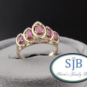 May include: A gold ring featuring five marquise-cut red gemstones arranged in a crown-like setting. The band is polished gold, and the stones are held in individual prong settings. The logo SJB Sherrie's Jewelry Box is visible in the corner.