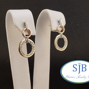 May include: A pair of gold and silver dangle earrings with a circle design. The circles are set with small clear stones. The earrings are on a white display stand.  SJB Sherrie's Jewelry Box.