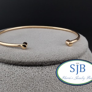 May include: A gold bangle bracelet with rounded ends. The bracelet is on a gray surface with a logo that reads "SJB Sherrie's Jewelry Box".