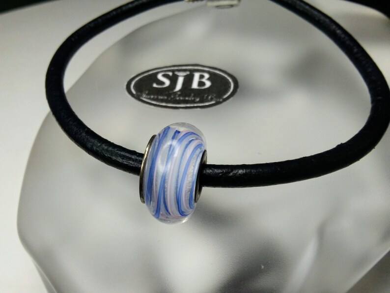 May include: A white glass bead with blue and pink swirls on a black leather bracelet. The bracelet has a silver charm with the text "SJB Jewelry Boutique" on it.