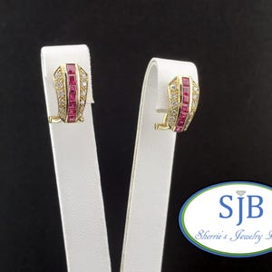 May include: A pair of gold-toned earrings with a rectangular ruby and diamond design. The earrings are displayed on a white stand against a black background. The earrings have a clip-on style.