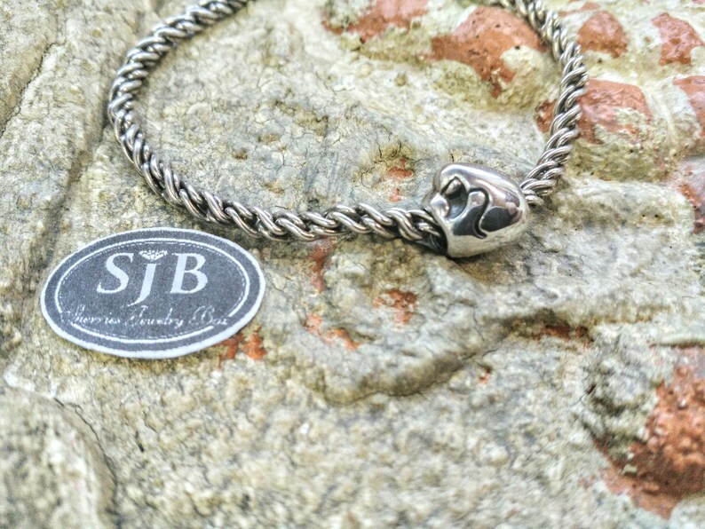 Pu&ograve; includere: Un braccialetto con ciondolo in argento con un ciondolo a forma di cuore che presenta due cuori intrecciati. Il ciondolo &egrave; attaccato a una catena in argento con un design a corda intrecciata. Il braccialetto &egrave; su una superficie grigia testurizzata. L'immagine include anche un'etichetta che recita "SJB Jewelry Box".