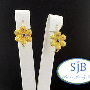 May include: A pair of gold-tone earrings featuring a five-petal flower design with intricate filigree wirework and a small blue stone at the centre of each blossom. The earrings are suspended from curved wire hooks and display a delicate, openwork pattern.