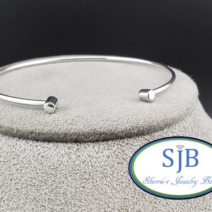 May include: A silver bangle bracelet with rounded ends. The bracelet is on a gray surface with a logo that says "SJB Sherrie's Jewelry Box".