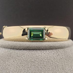 Może przedstawiać: Złota bransoletka typu bangle z jednym prostokątnym szmaragdowo zielonym kamieniem osadzonym w środku.